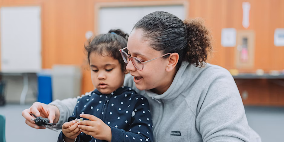 An adult and a young child sit together indoors. As they work with a small object, the adult, wearing glasses and a light gray sweater, guides the child in her navy blue polka-dot hoodie through her My First IEP activities. Both are focused and engaged.