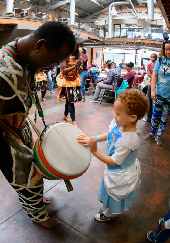 Man with an African drum leans down as a young child beats the drum